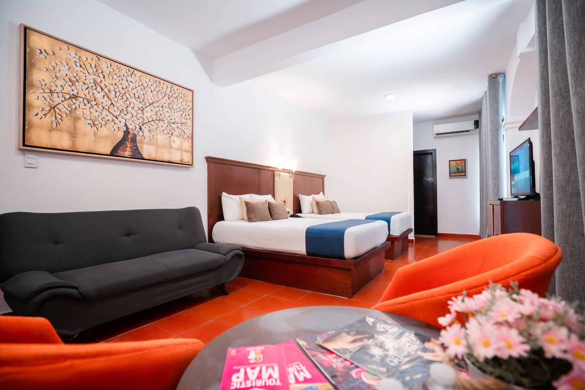 Habitación de hotel con dos camas, sofá gris, sillas naranjas y arte en la pared.