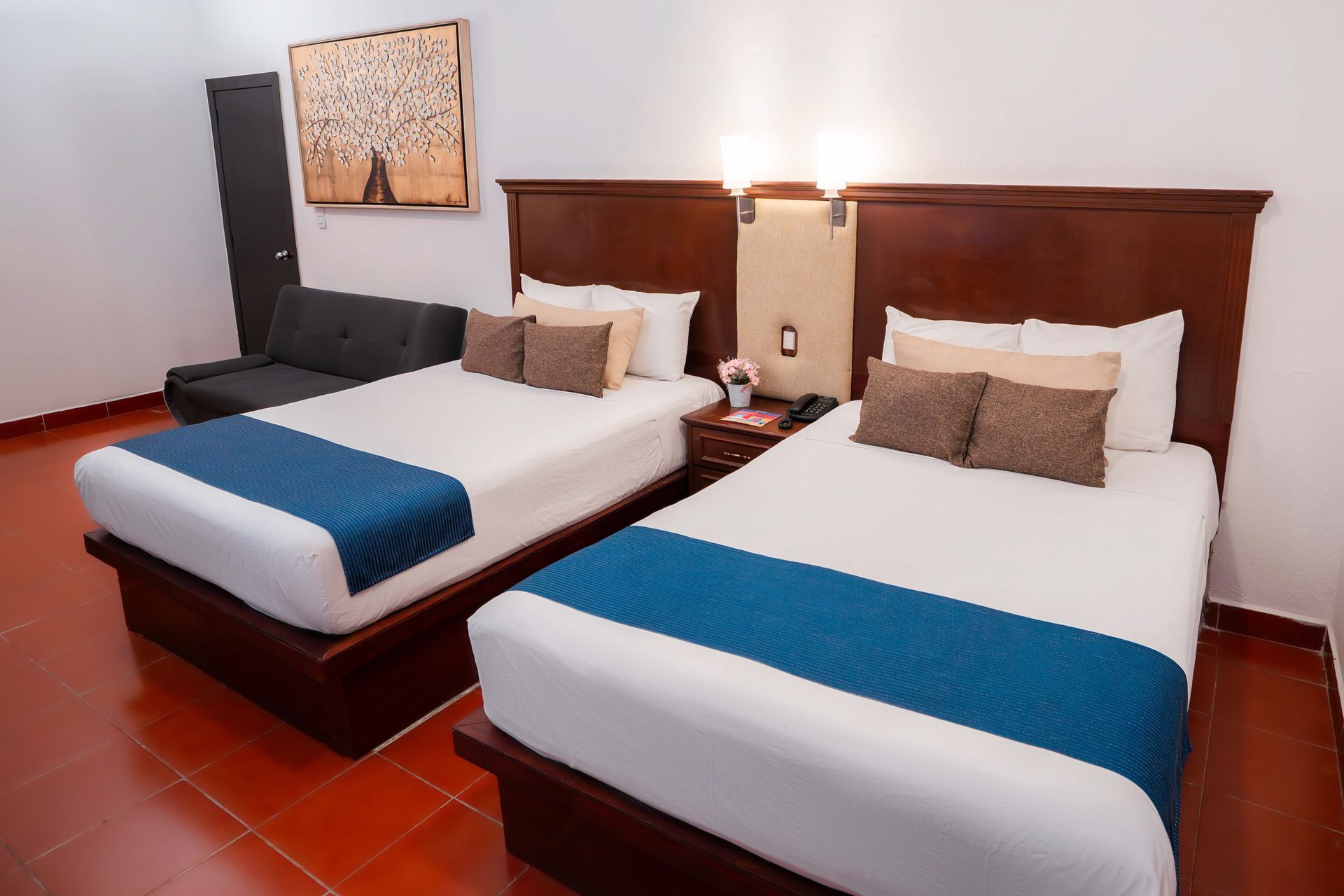 Dos camas con sábanas blancas y corredores azules, cabeceros de madera oscura y un pequeño sofá en una habitación de hotel.
