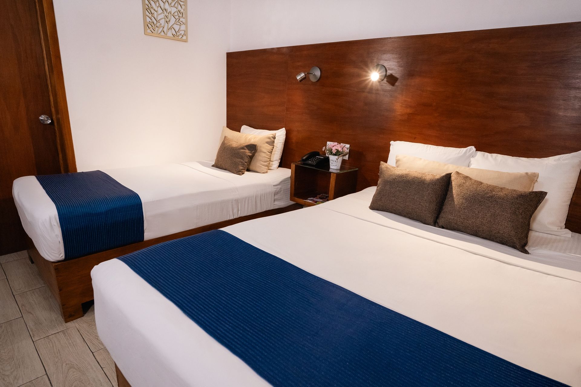 Habitación de hotel con dos camas, sábanas blancas, detalles en azul, cabecera de madera oscura, mesita de noche pequeña.