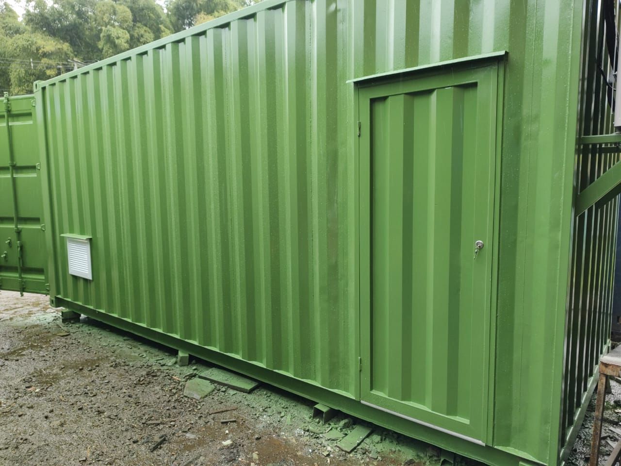 Contenedor de transporte verde con puerta, colocado sobre grava al aire libre entre árboles.