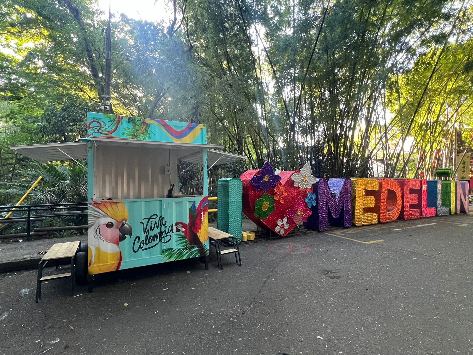 Un colorido letrero de arte callejero con la palabra “MEDELLÍN” junto a un pequeño quiosco en un parque soleado.