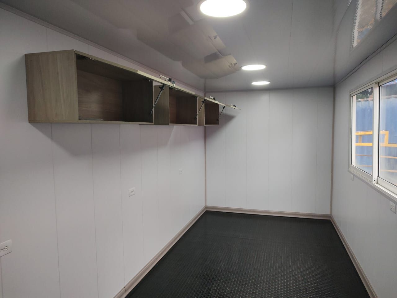 Habitación prefabricada vacía con paredes grises, luces en el techo, armarios de pared y una ventana a la derecha.