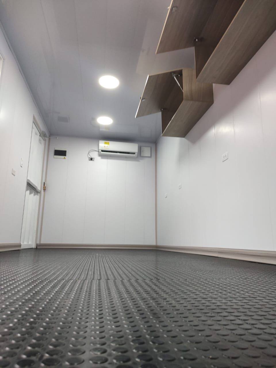 Habitación blanca vacía con suelo texturizado gris, luces de techo, aire acondicionado mural y estantes de madera.
