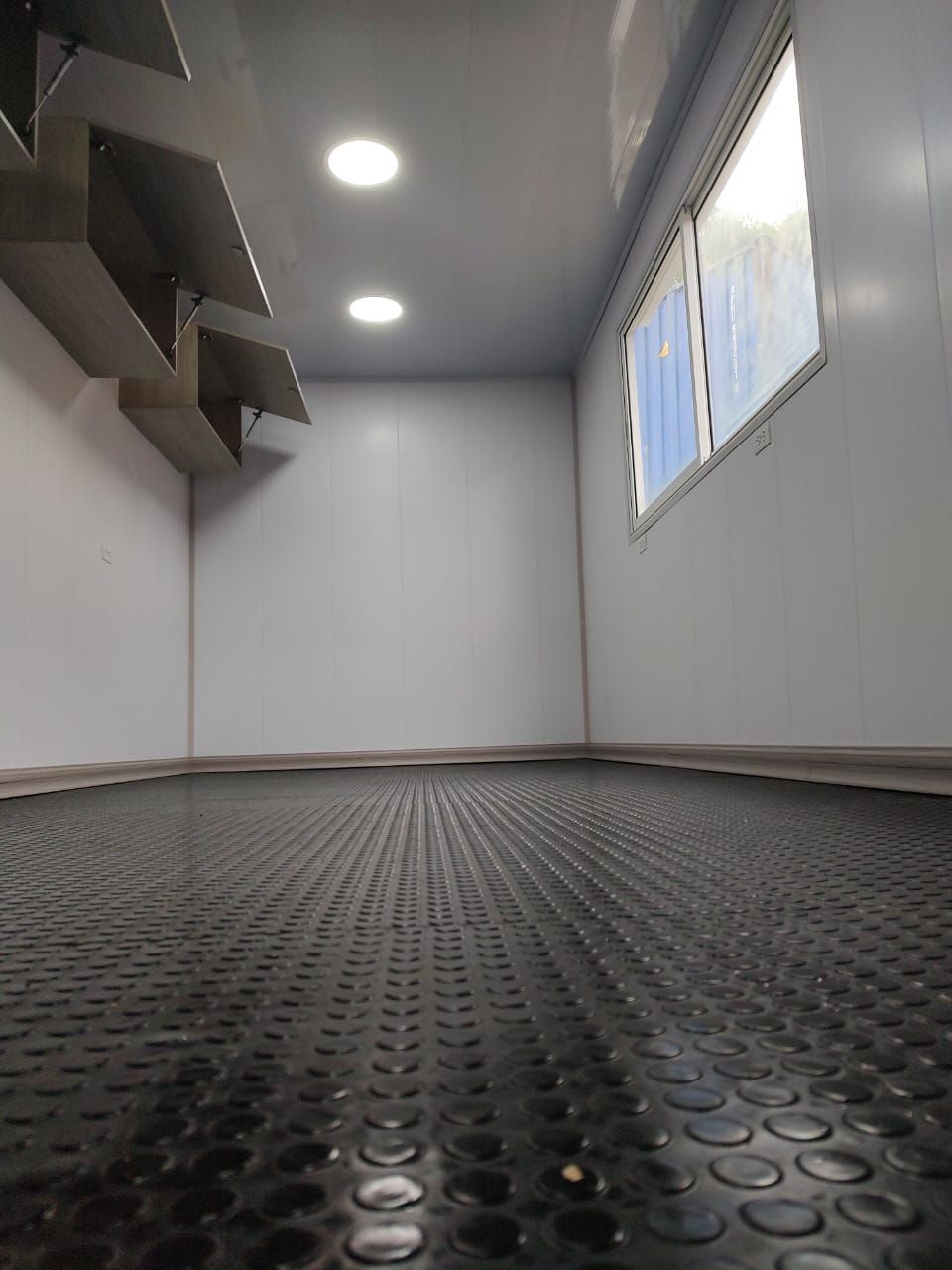 Habitación gris vacía con esteras de goma negras en el suelo, luces en el techo y una ventana a la derecha.