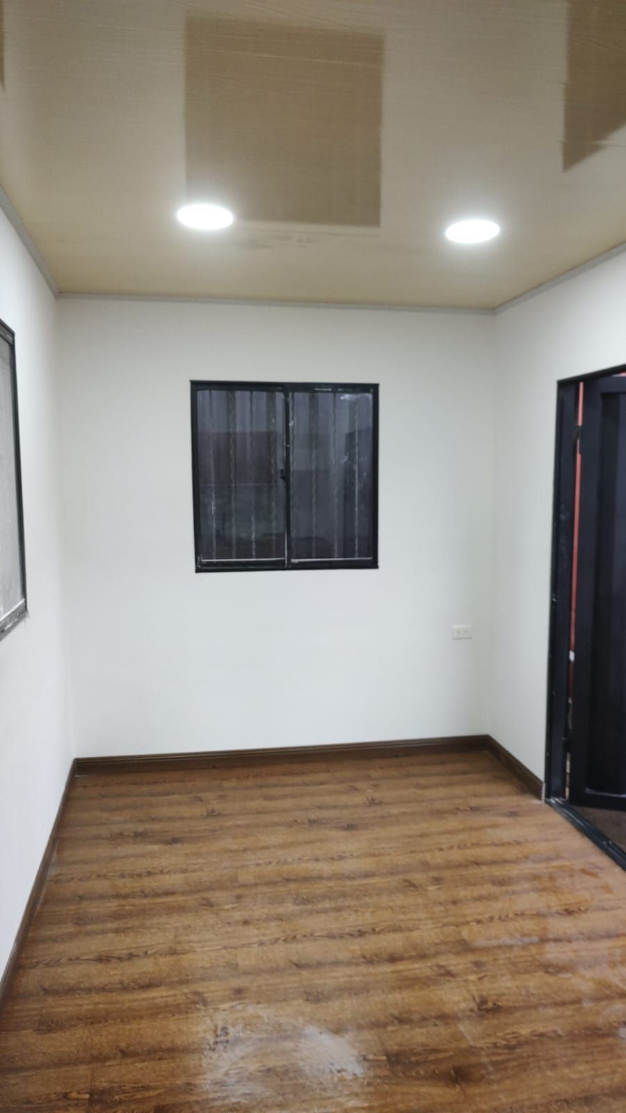Habitación vacía con suelo de madera, paredes blancas, luces en el techo y una ventana en la pared del fondo.