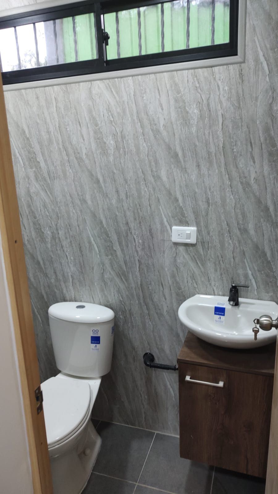 Baño pequeño con inodoro, lavabo mural, azulejos grises y una ventana esmerilada en la parte superior.