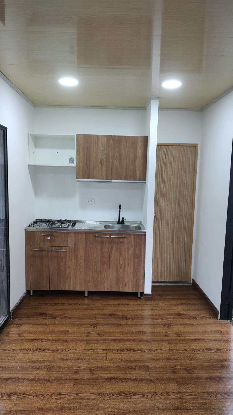 Pequeña cocina con armarios de madera, fregadero, fogones y puerta cerrada sobre suelo de madera.