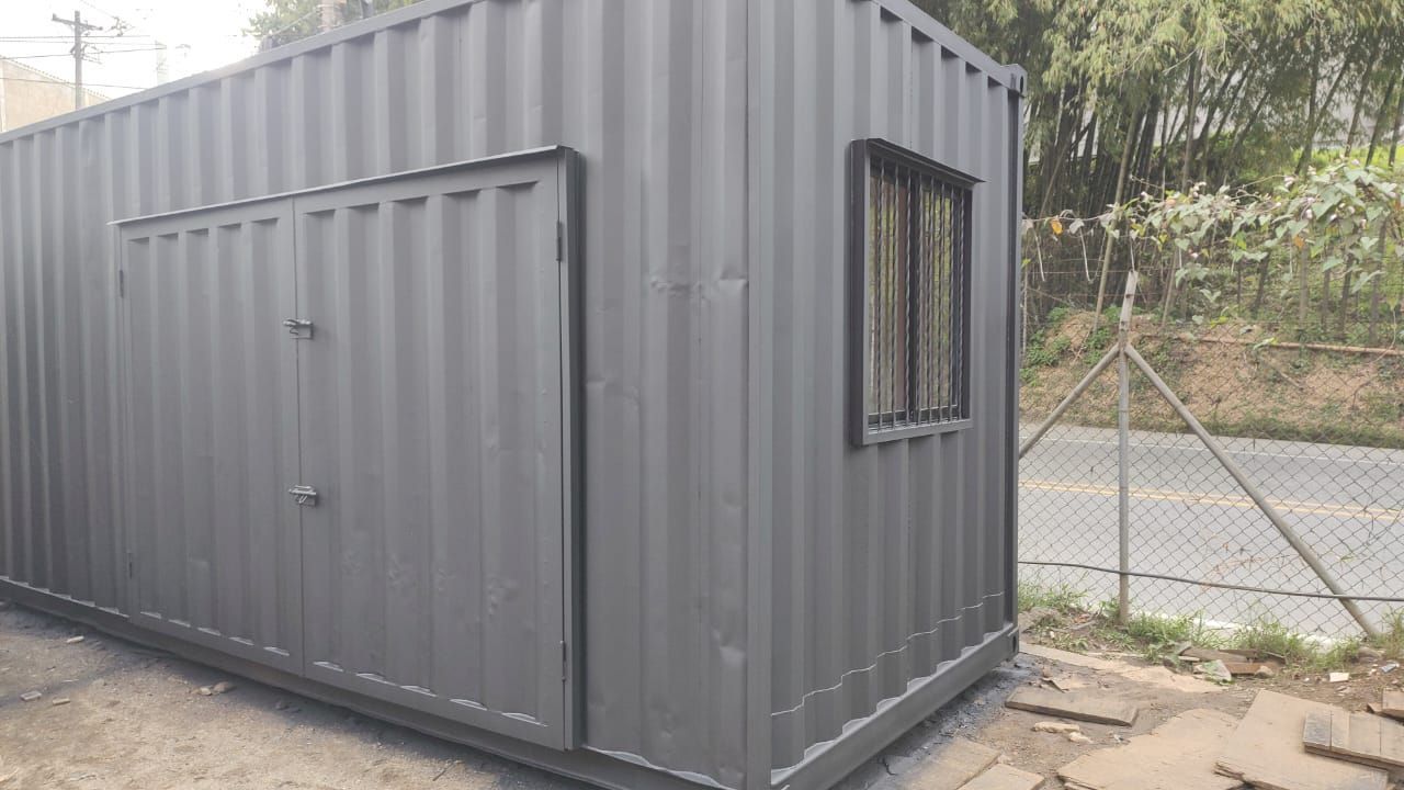Contenedor de transporte gris con una pequeña ventana, situado al aire libre junto a una valla y una carretera.