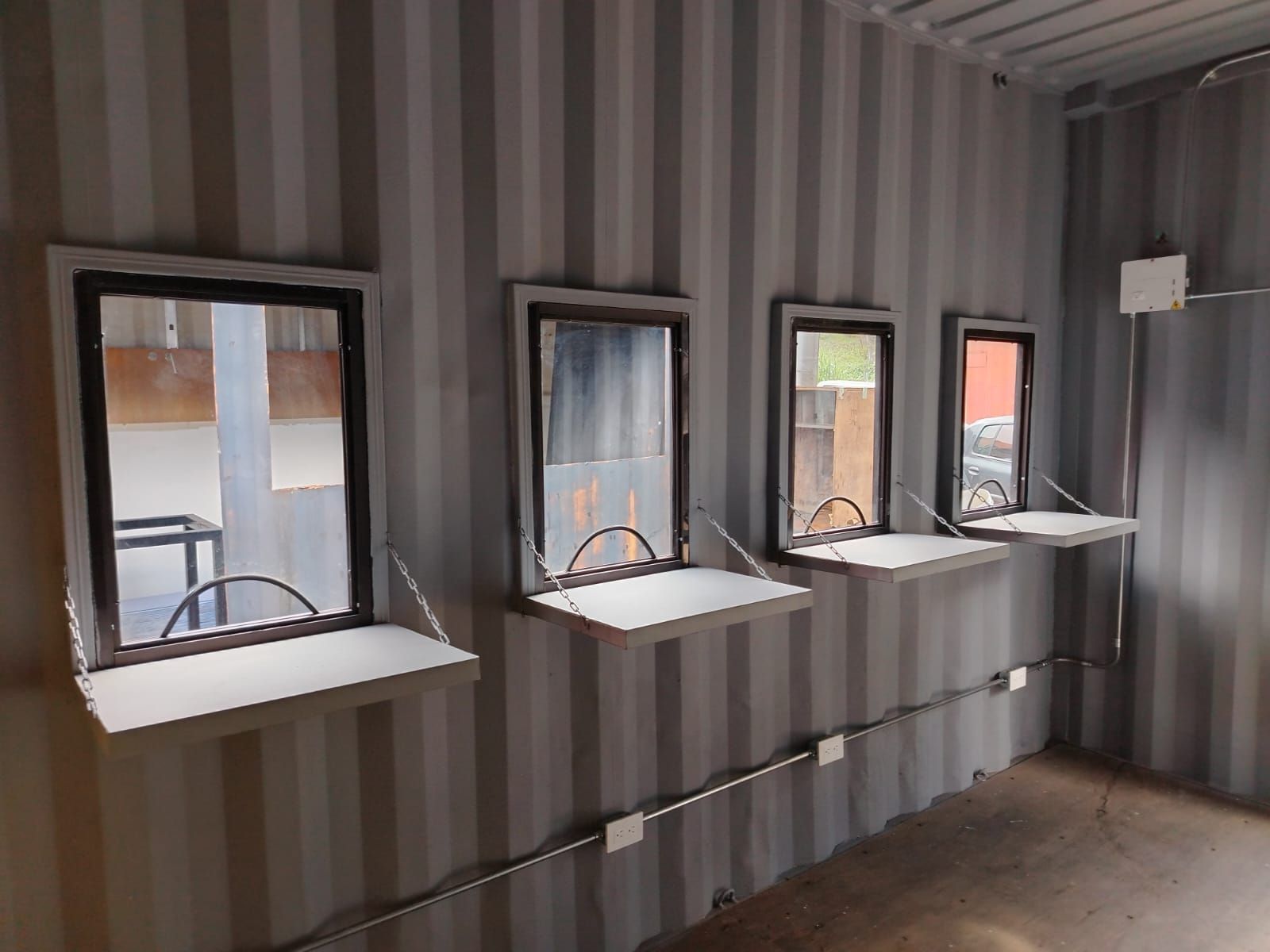 Interior de un contenedor de transporte con cuatro pequeñas ventanas y estantes blancos en una habitación gris a rayas.