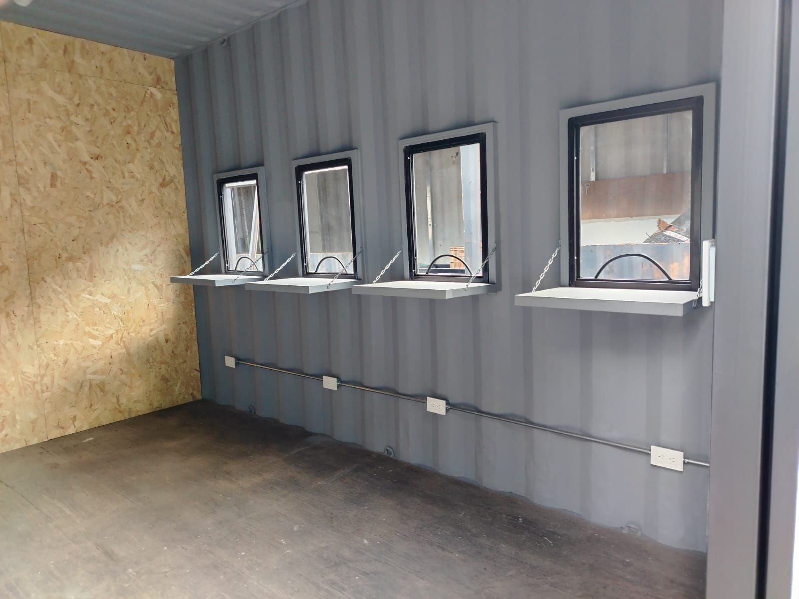 Habitación industrial vacía con paredes de chapa ondulada gris, ventanas pequeñas y una sección de pared revestida de madera