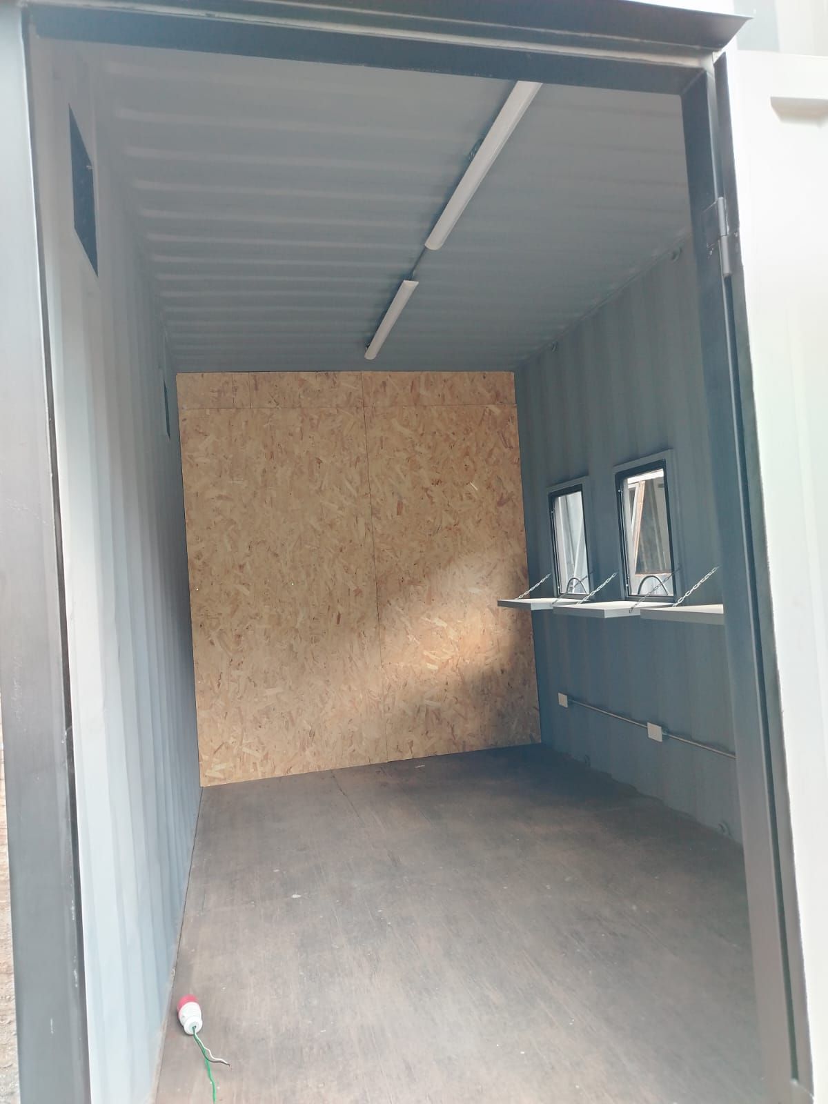 Contenedor de transporte gris vacío con pared de madera contrachapada, ventanas laterales y suelo de hormigón.