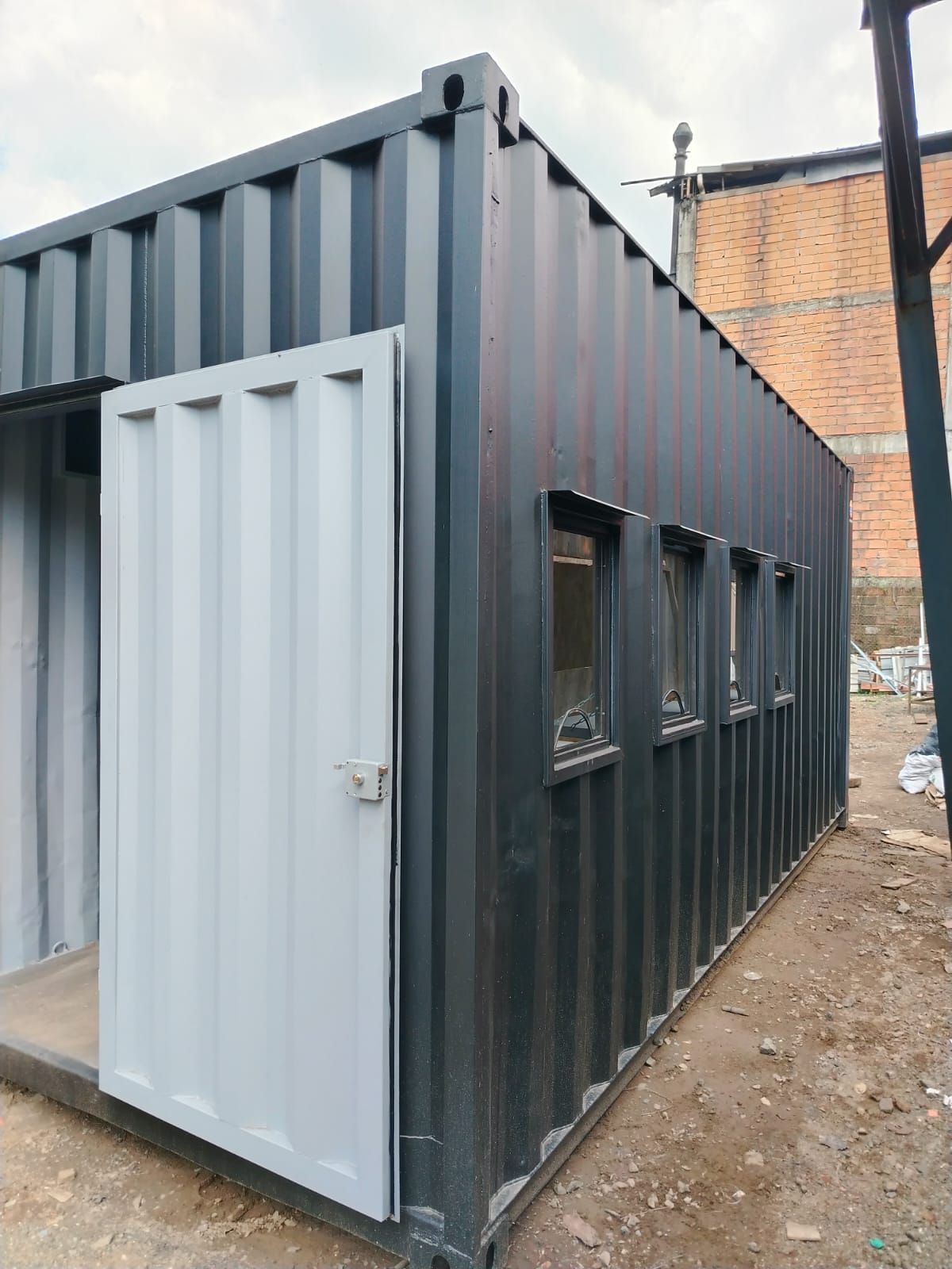 Oficina construida con un contenedor de transporte de color gris oscuro, con puerta blanca y ventanas pequeñas
