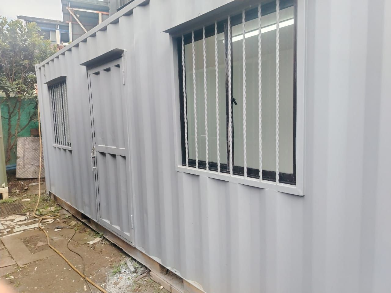 Oficina portátil de metal corrugado gris con ventanas enrejadas a lo largo de una estrecha pasarela exterior.