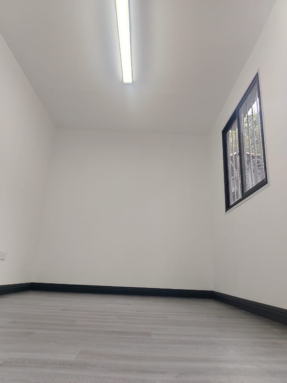 Habitación blanca vacía con suelo gris, lámpara de techo y una pequeña ventana a la derecha.