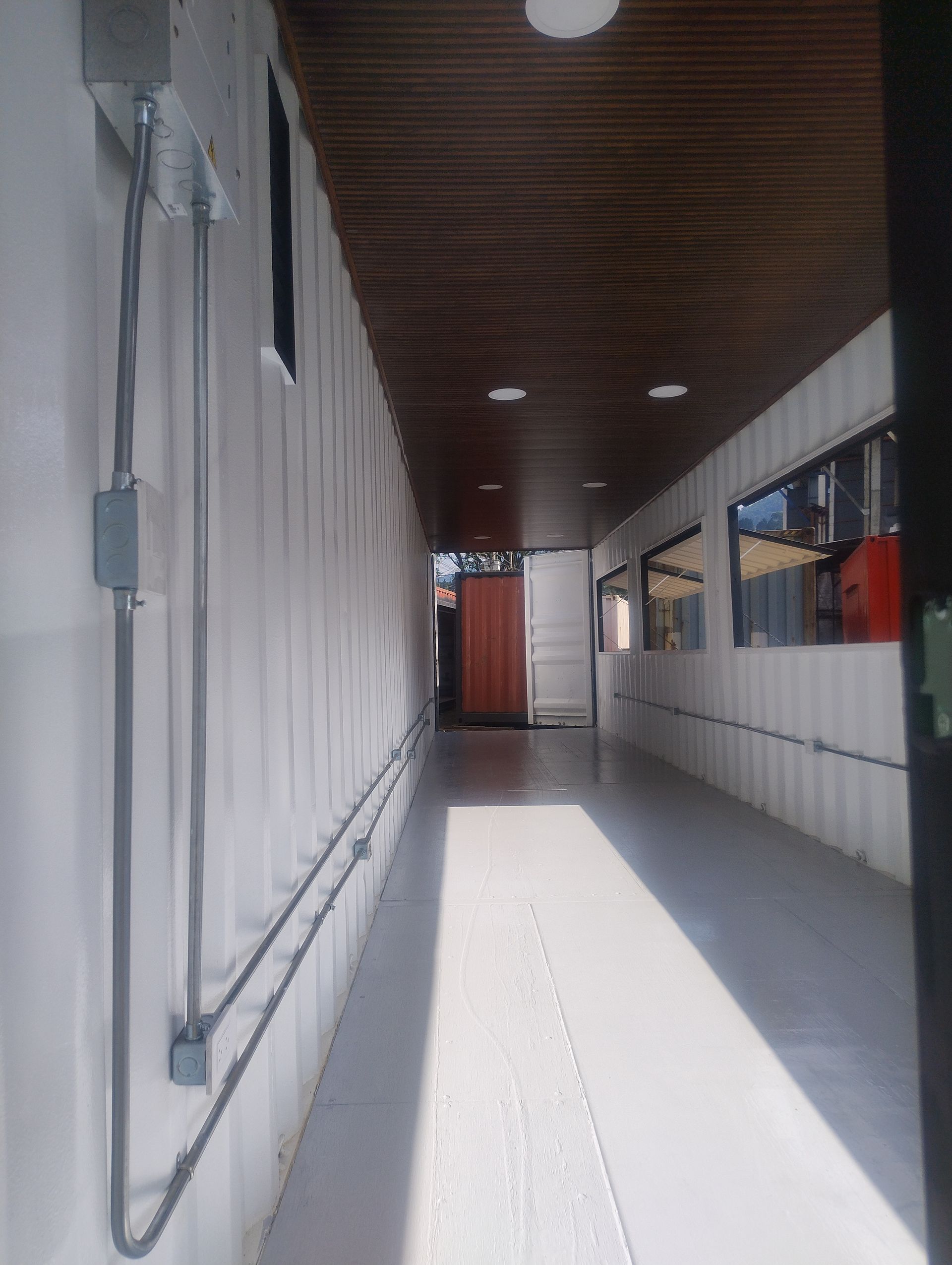 Estrecho pasillo exterior con paredes blancas, techo de madera y luz solar que proyecta una brillante sombra diagonal.