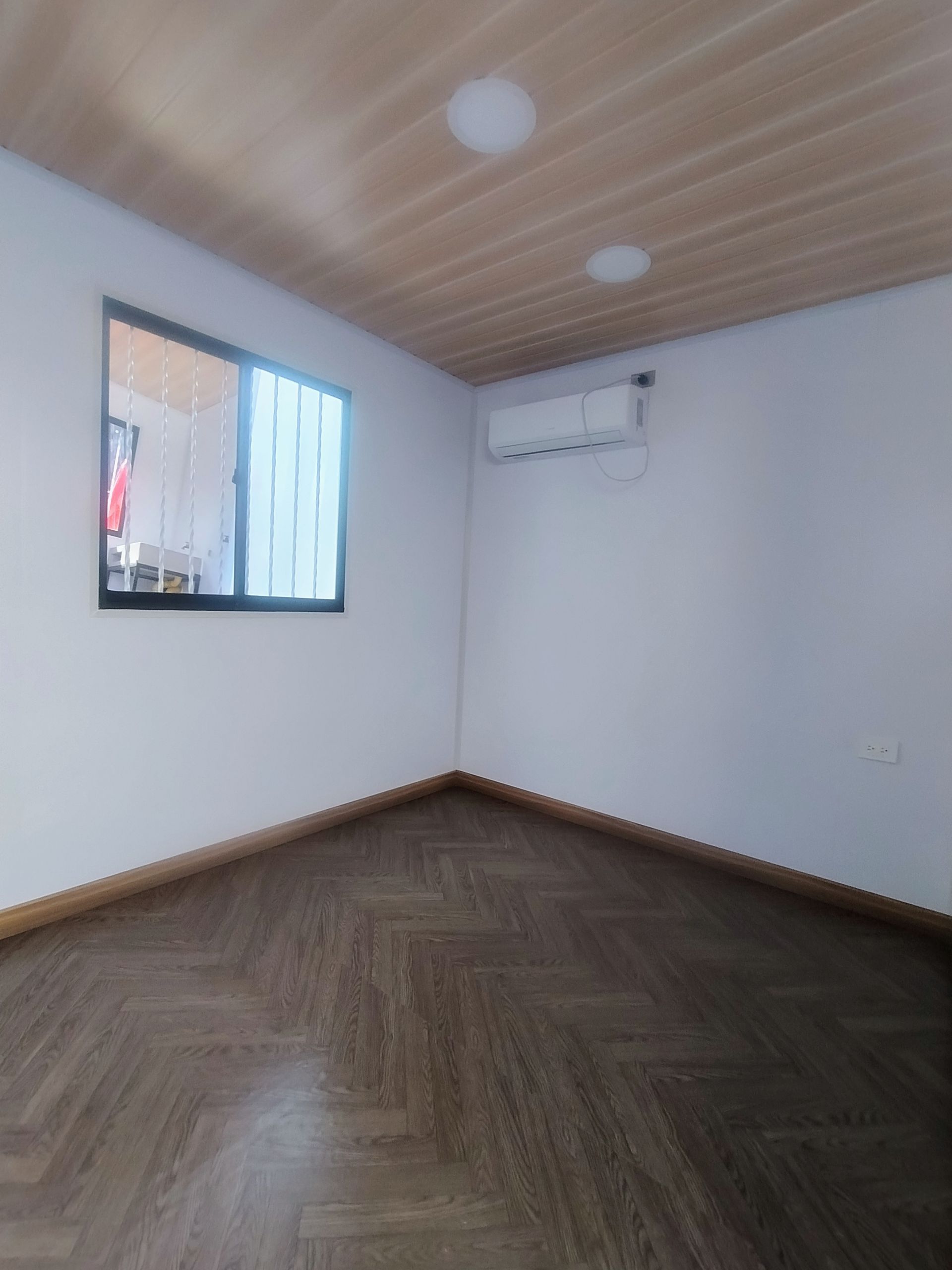 Habitación vacía con suelo de madera, paredes blancas, luces de techo y aire acondicionado de ventana.