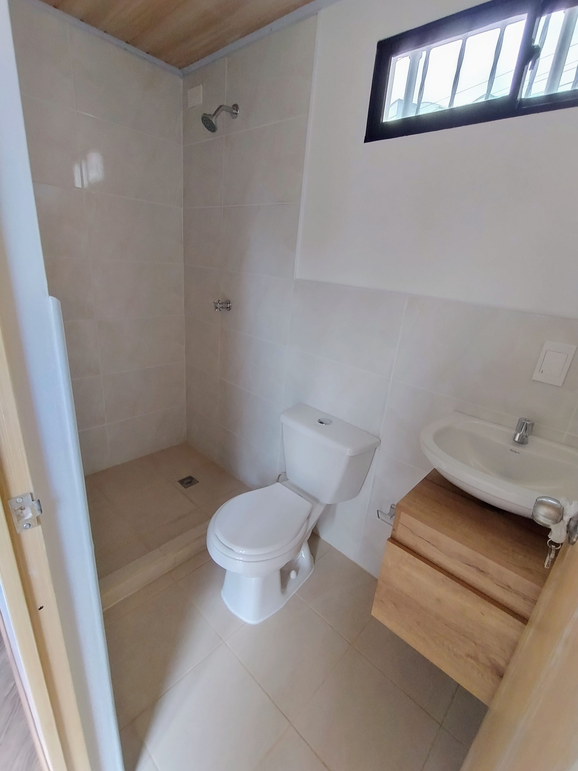 Baño pequeño con inodoro, lavabo, ducha y ventana alta, acabado en azulejos blancos y madera clara.