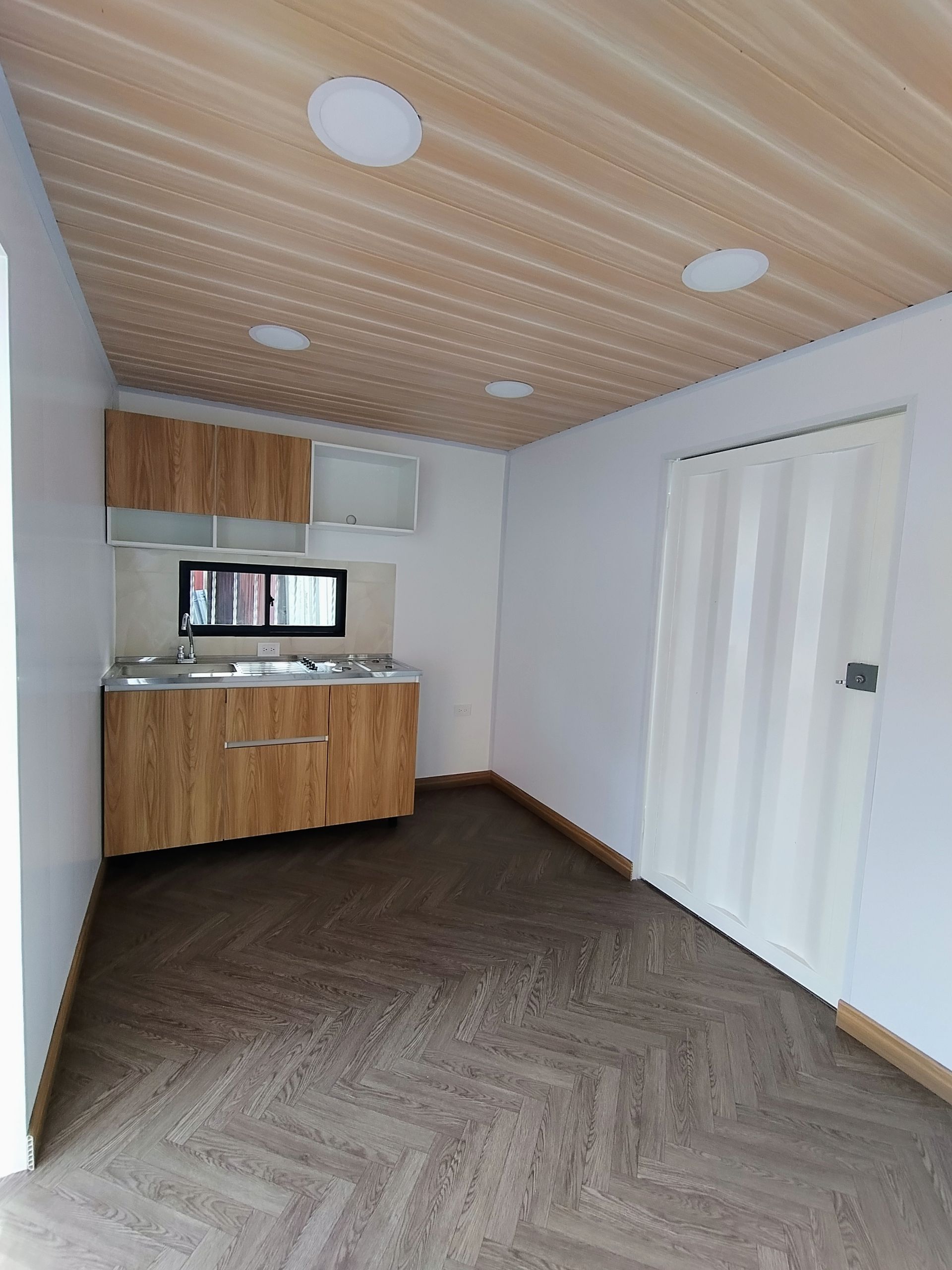 Cocina o cocineta moderna vacía con armarios de madera, suelo de espiga y puertas plegables blancas.