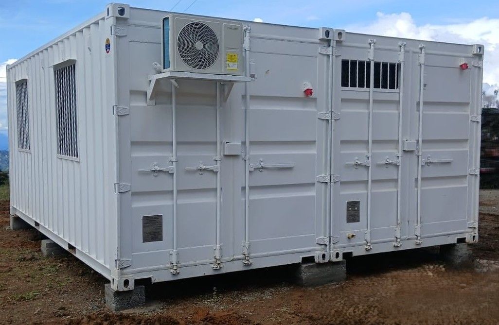 Contenedor de transporte blanco con ventanas enrejadas y una unidad de aire acondicionado, ubicado en un terreno de grava.