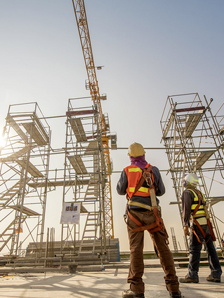 Two construction workers are standing in front of a crane on a construction site.