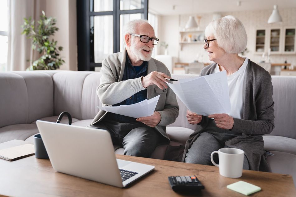 Two people reviewing paperwork on a sofa beside a laptop in a bright living room