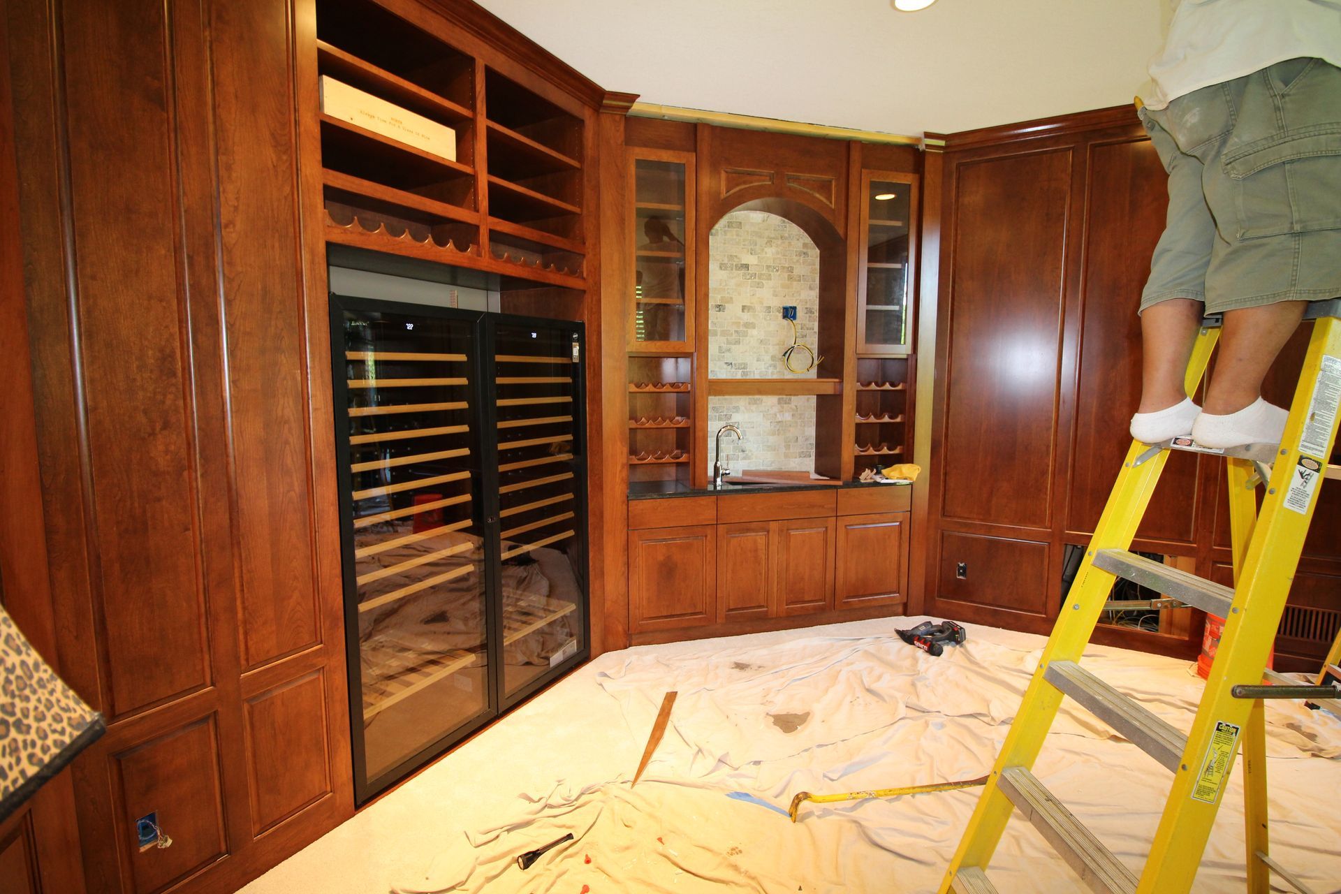 Built-in wine closet with wood cabinets and a person on a ladder working on the ceiling