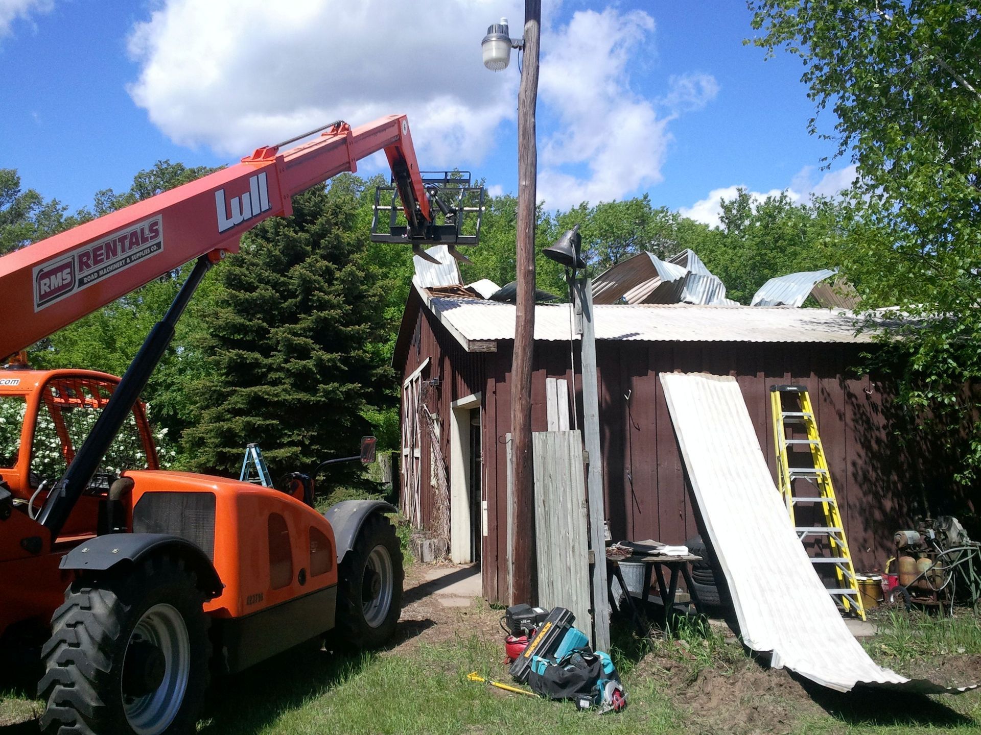 Orange excavator with raised arm beside a small shed roof under repair, with ladders and debris nearby