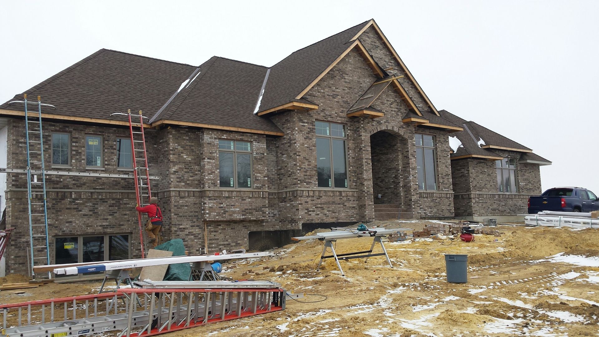 Stone house under construction with roof framing, ladders, and snowy ground