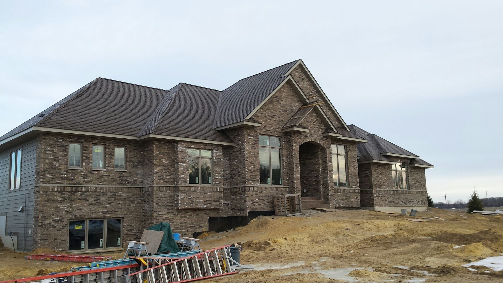Large stone house under construction on a dirt lot, with roof shingles and construction materials nearby