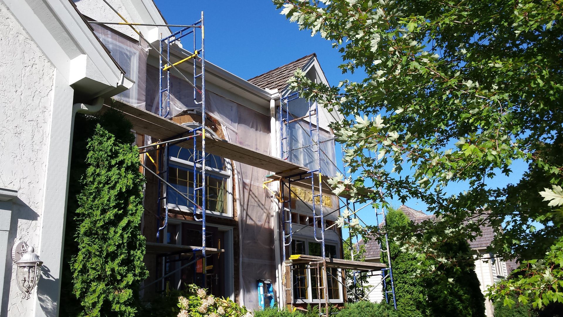 Two-story house with scaffolding and ladder under construction, viewed beside a tree on a sunny day