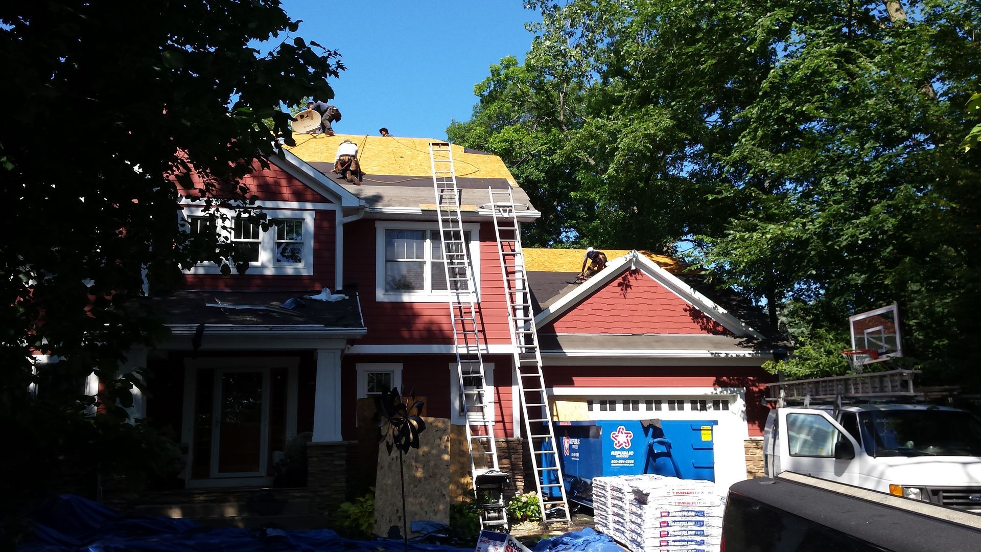 House with workers on roof and ladder, red siding, blue tarp, and truck in driveway