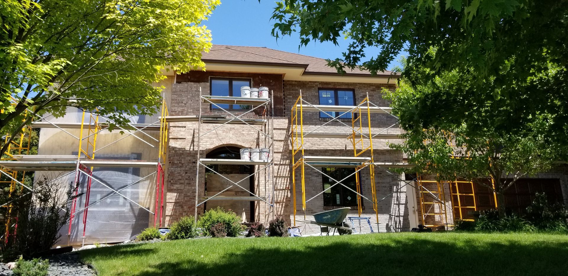 Two-story house under renovation with scaffolding and white tarp, surrounded by green trees and lawn