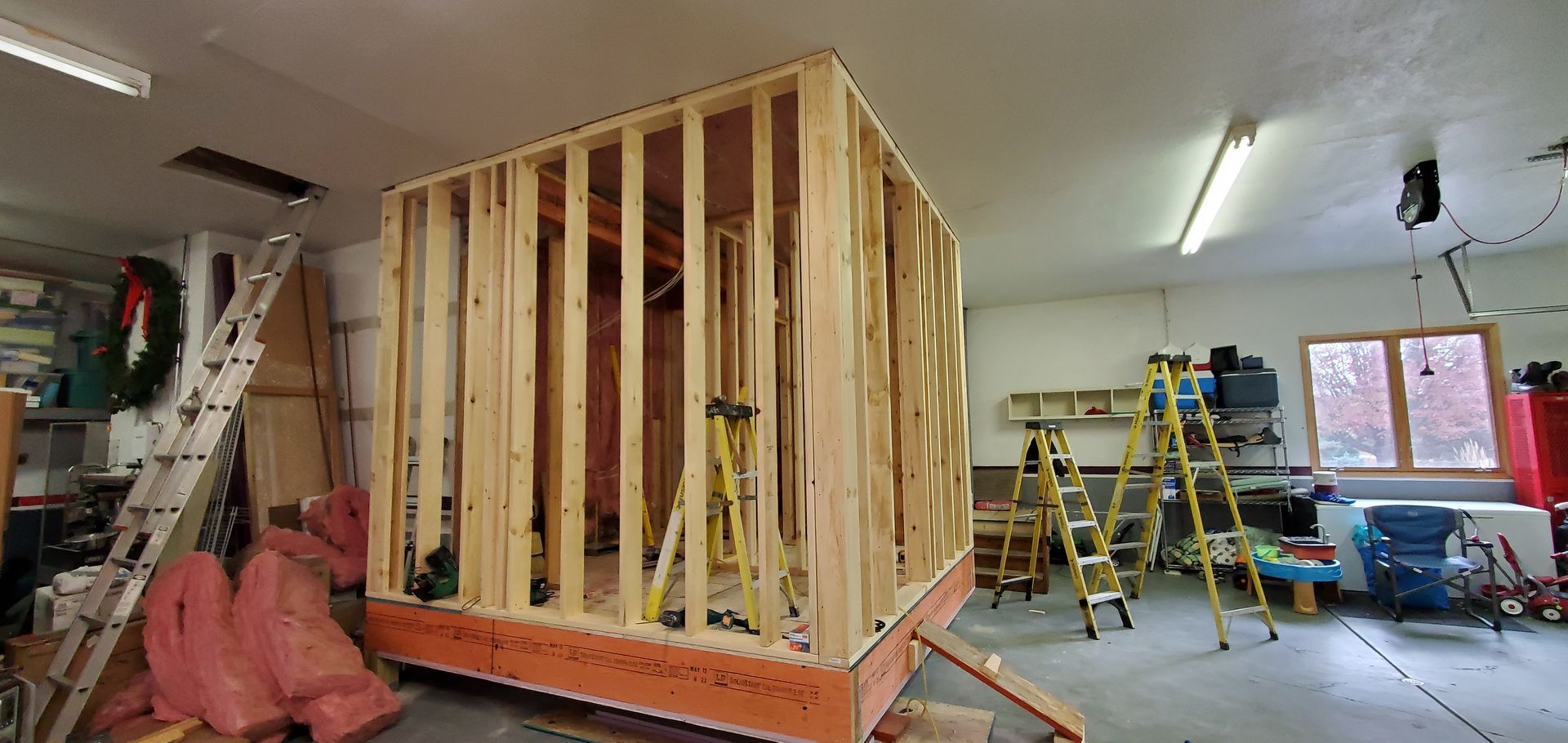 Framed wooden room under construction in a garage, with ladders, tools, and a workbench nearby