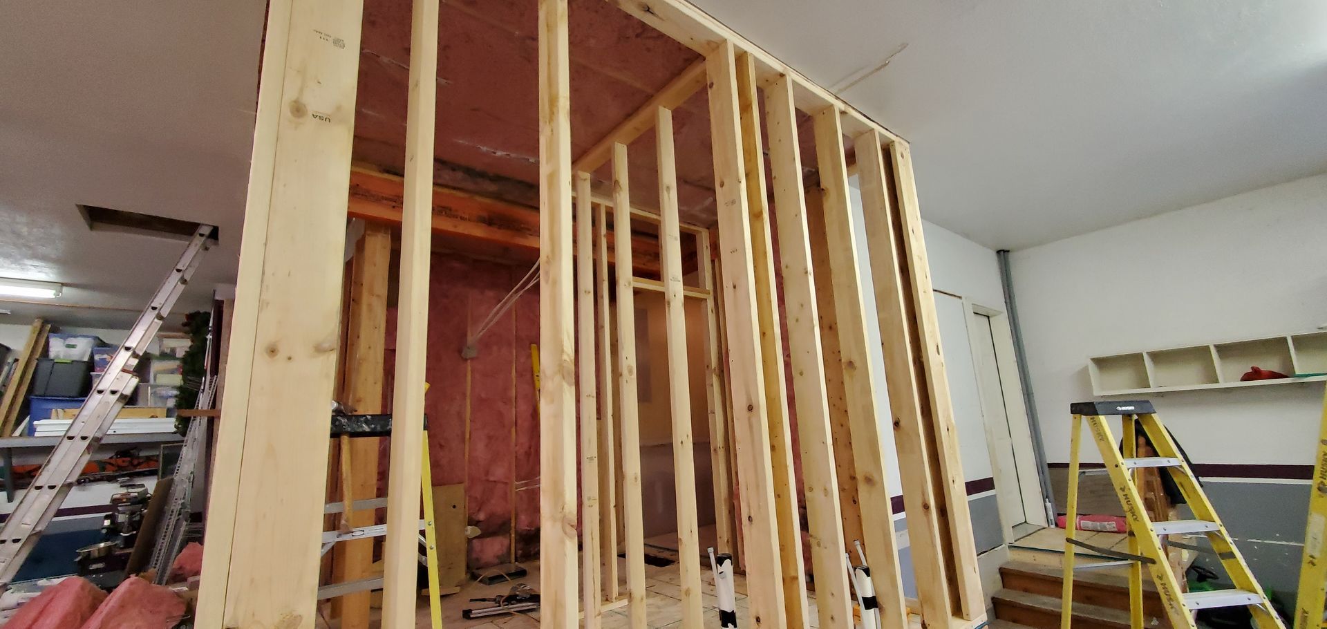 Framed interior wall under construction in a garage, with exposed wood studs and a step ladder nearby