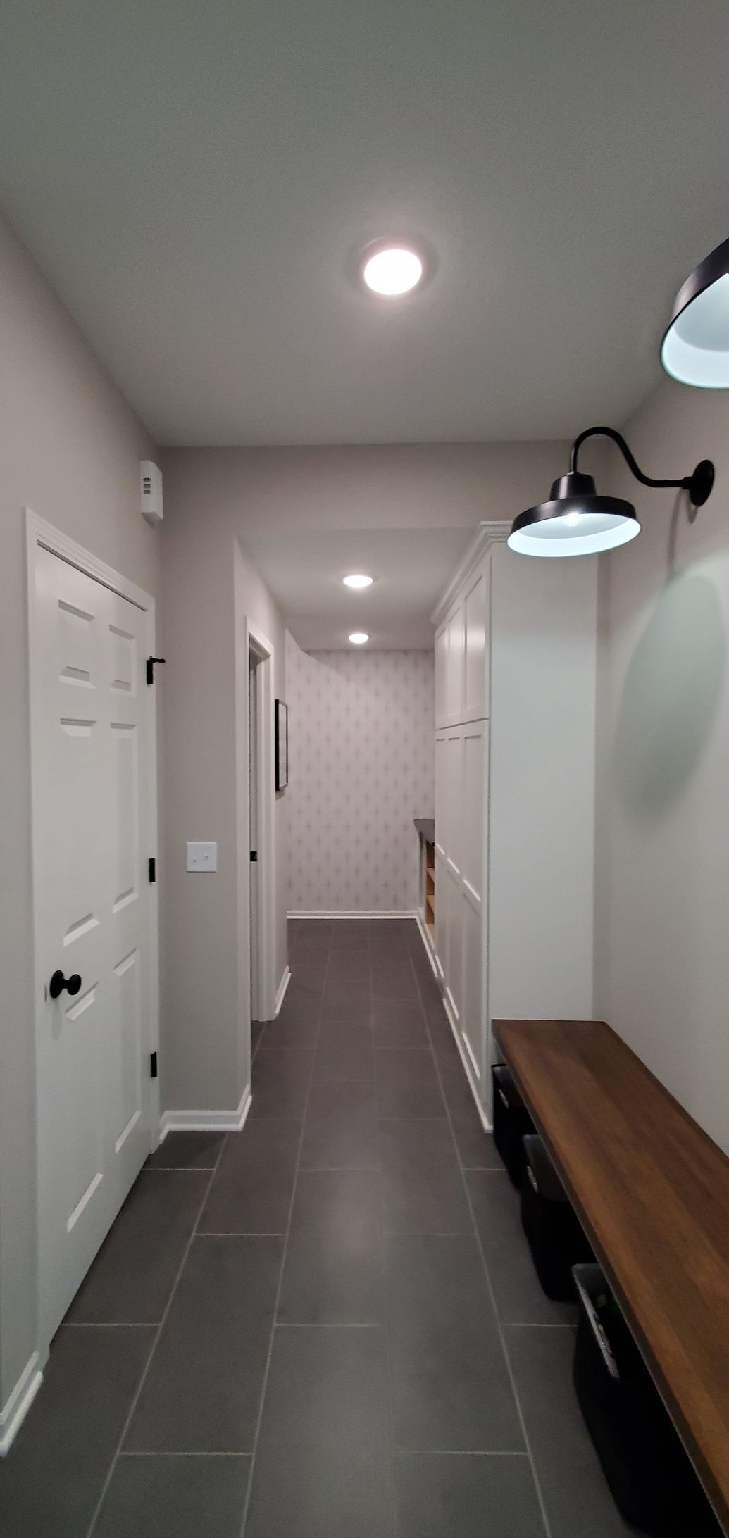Narrow hallway with gray tile floor, white walls, ceiling light, and a wooden bench along the right wall.