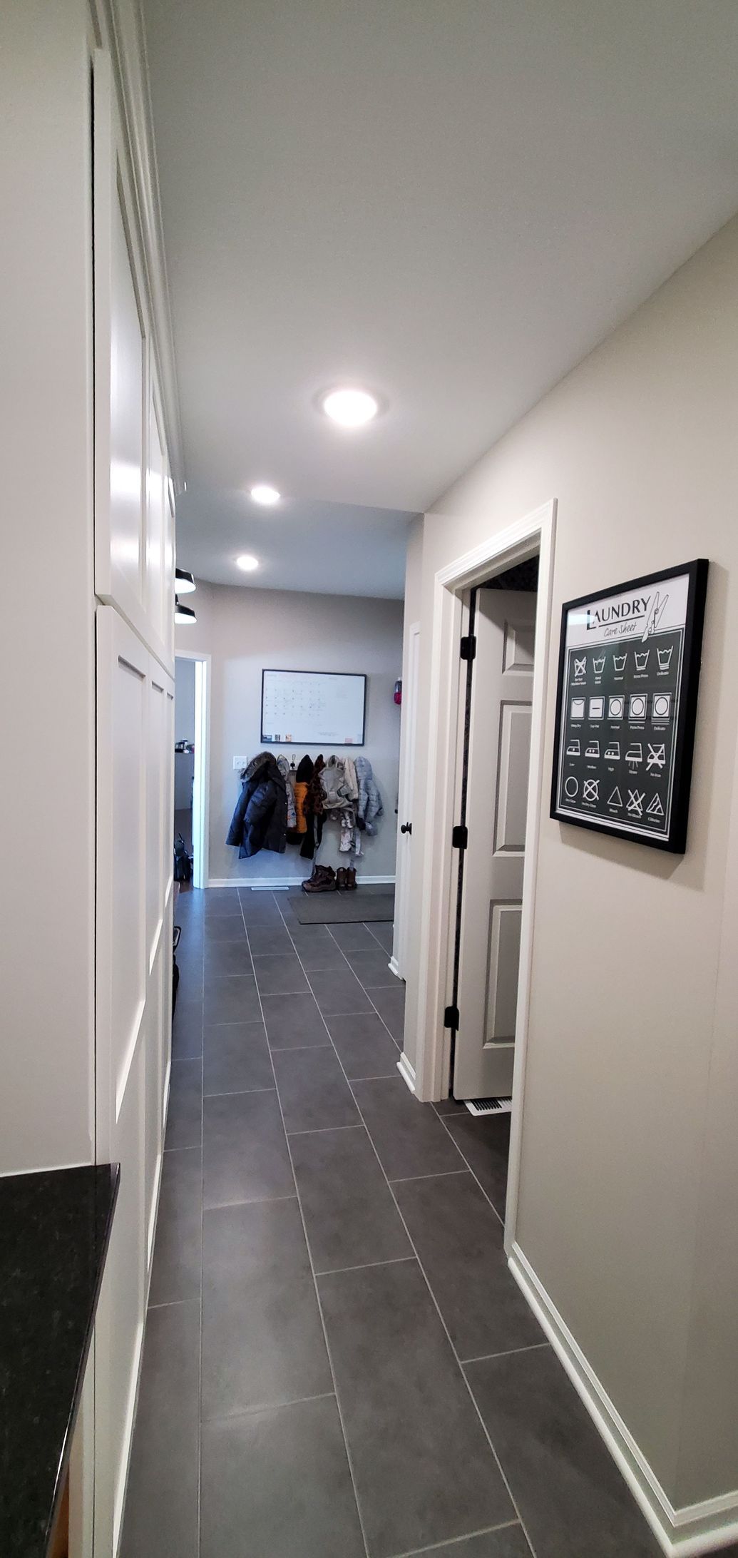 Narrow hallway with gray carpet, white walls, recessed lights, and a distant room with clothes hanging.