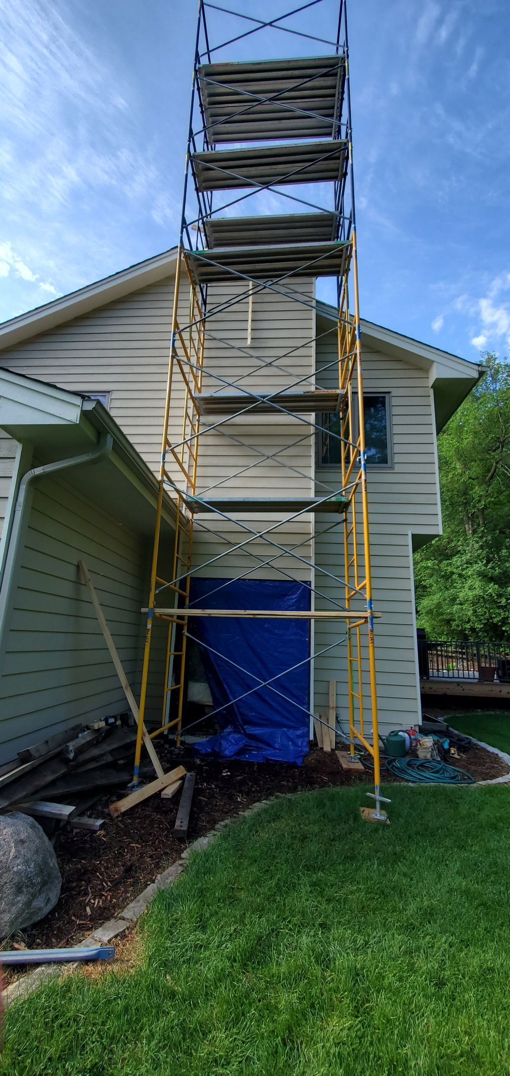 Scaffolding around a house exterior with a blue tarp at the base and ladder frames above
