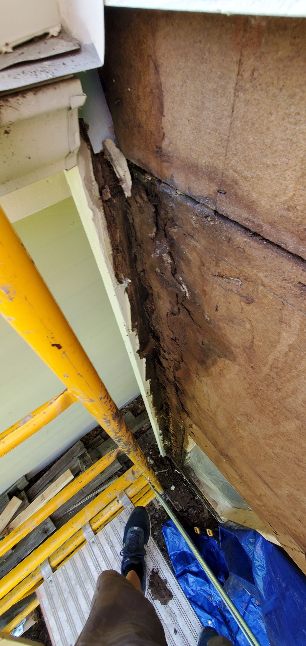 Close-up of a damaged wall seam with peeling material, exposed wood, and construction equipment below