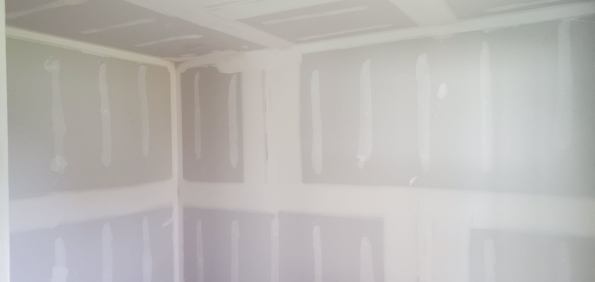 Unfinished drywall in a room, with taped seams and patches on white walls and ceiling.
