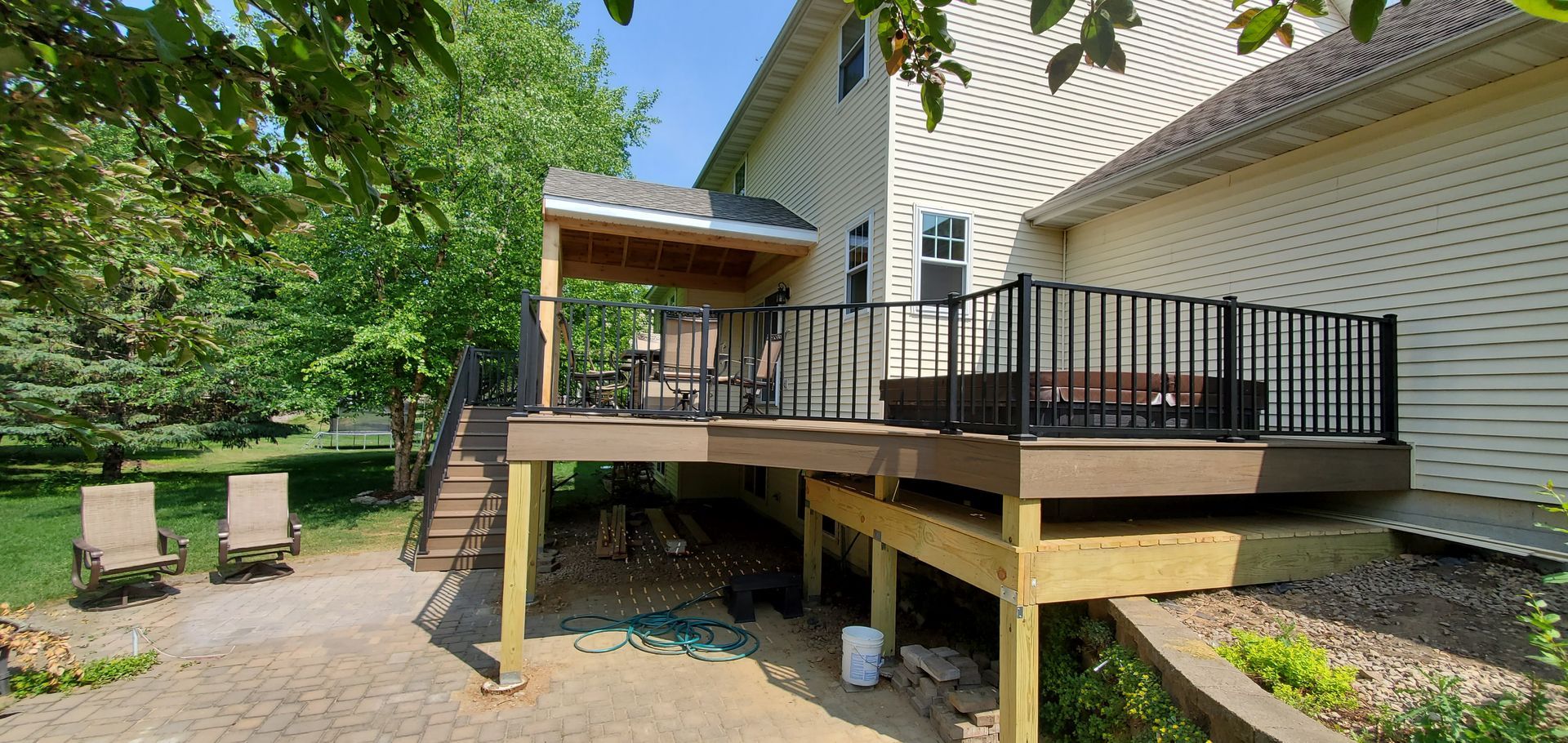 Raised wooden deck with black railing beside a white house, overlooking a stone patio and green yard