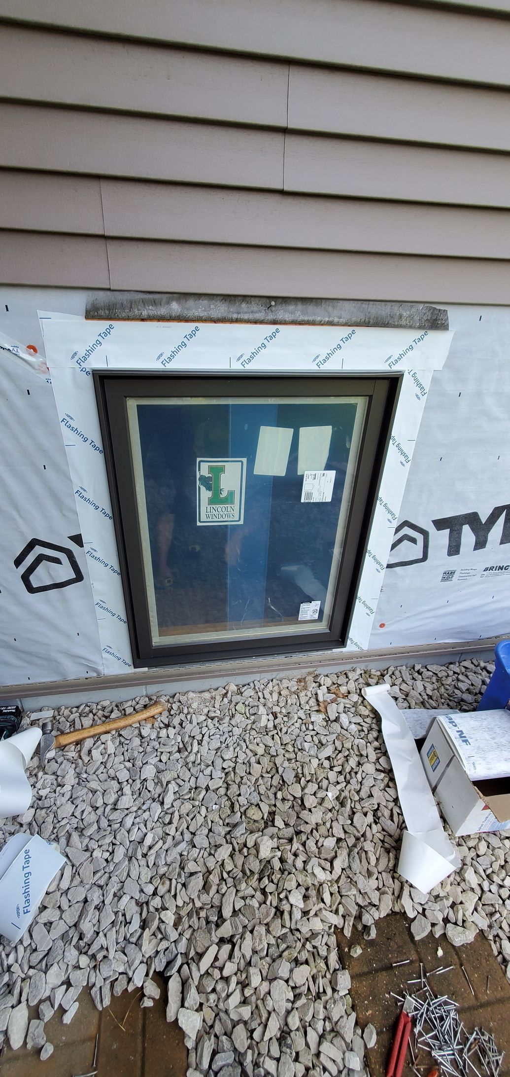 Basement window well under white siding, bordered by gravel and insulation scraps