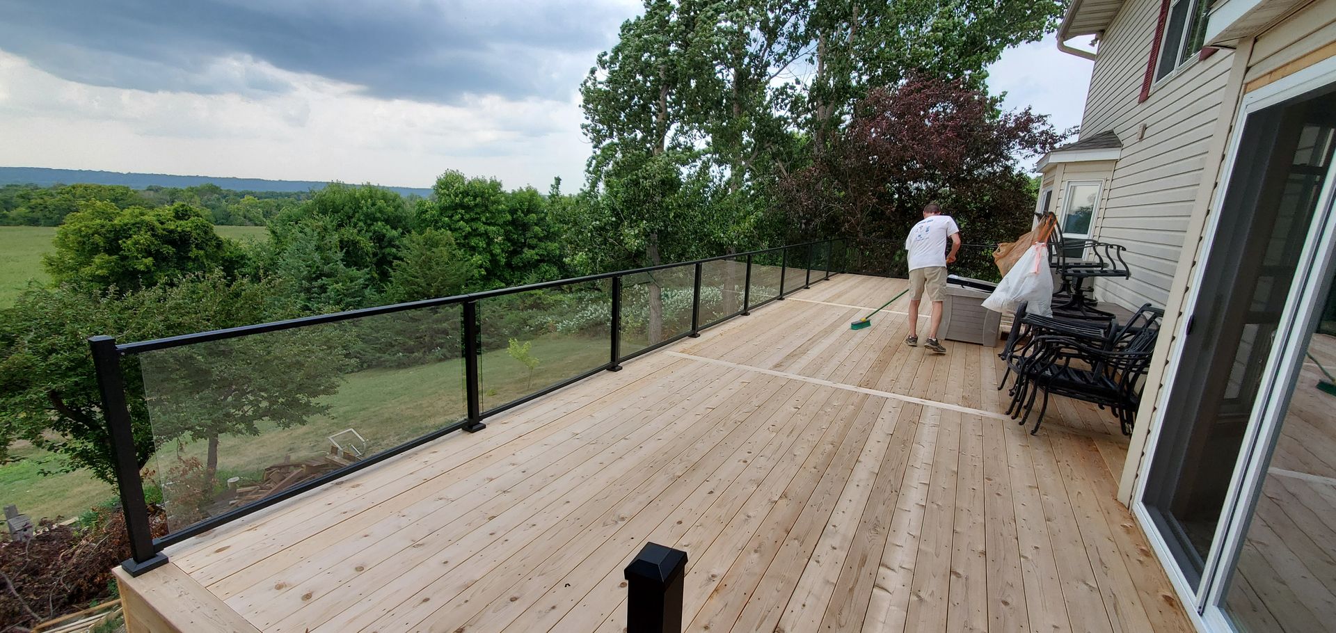 Wide wooden deck overlooking green hills, with two people near a house and outdoor seating.