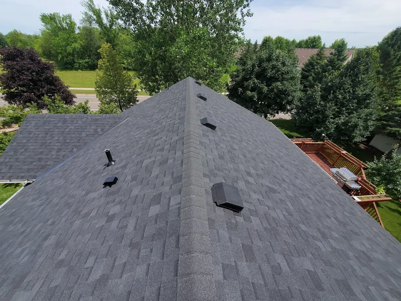 Dark gray shingled roof with vents, viewed from above against trees and neighboring houses