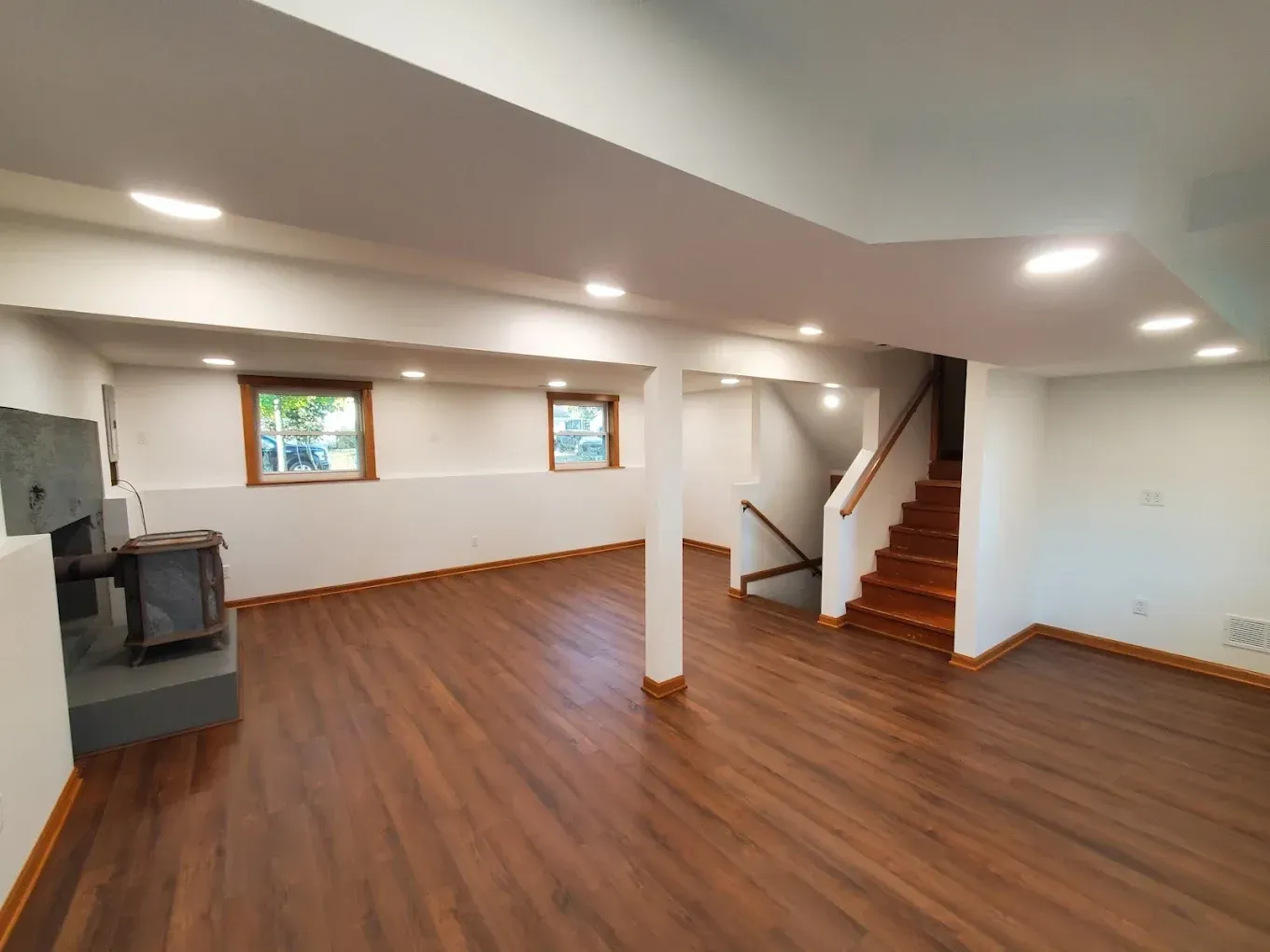 Empty basement living area with wood floor, white walls, fireplace, windows, and stairs to the upper level