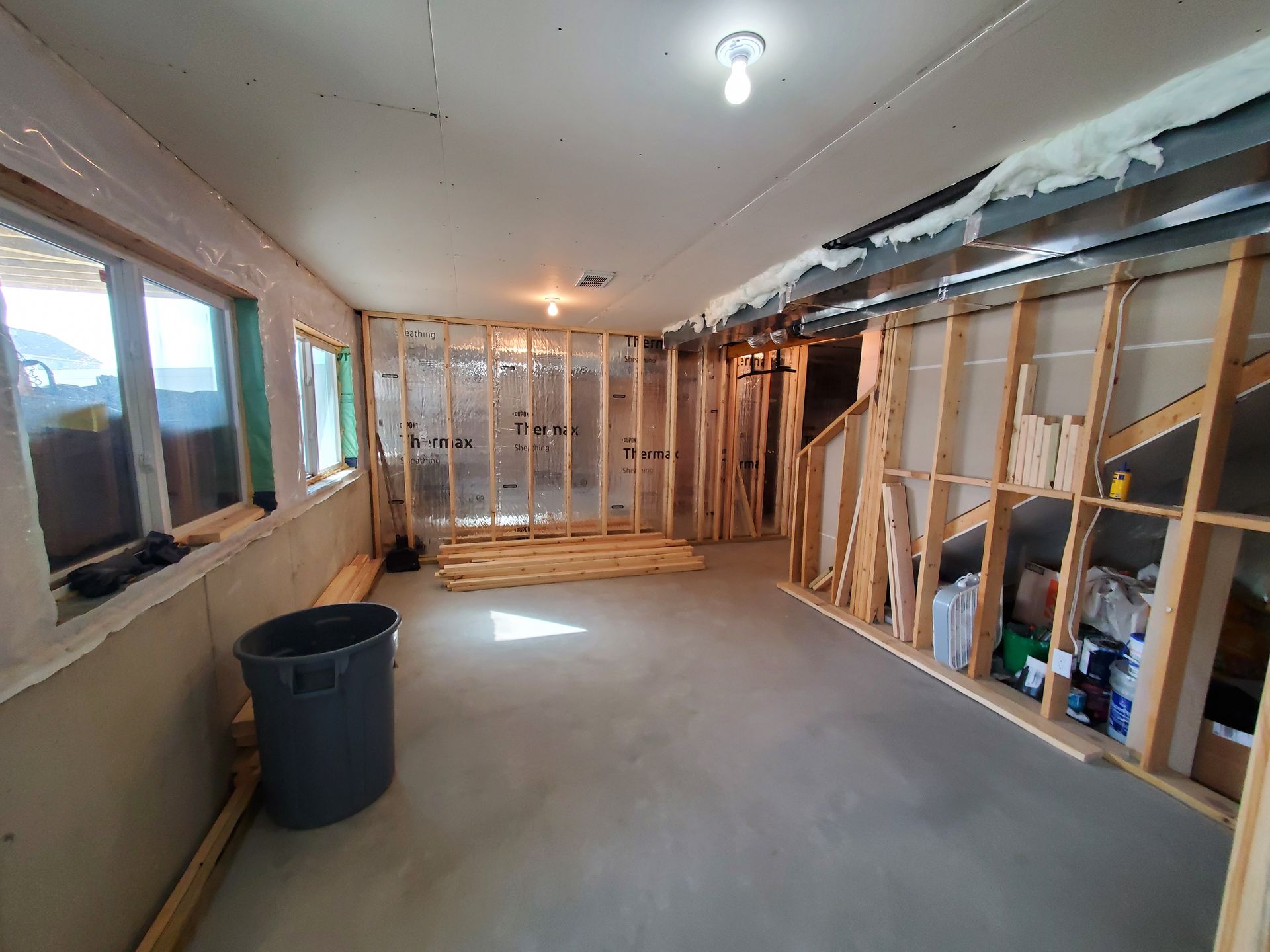 Partially renovated room with exposed studs, windows, and a trash bin on a concrete floor