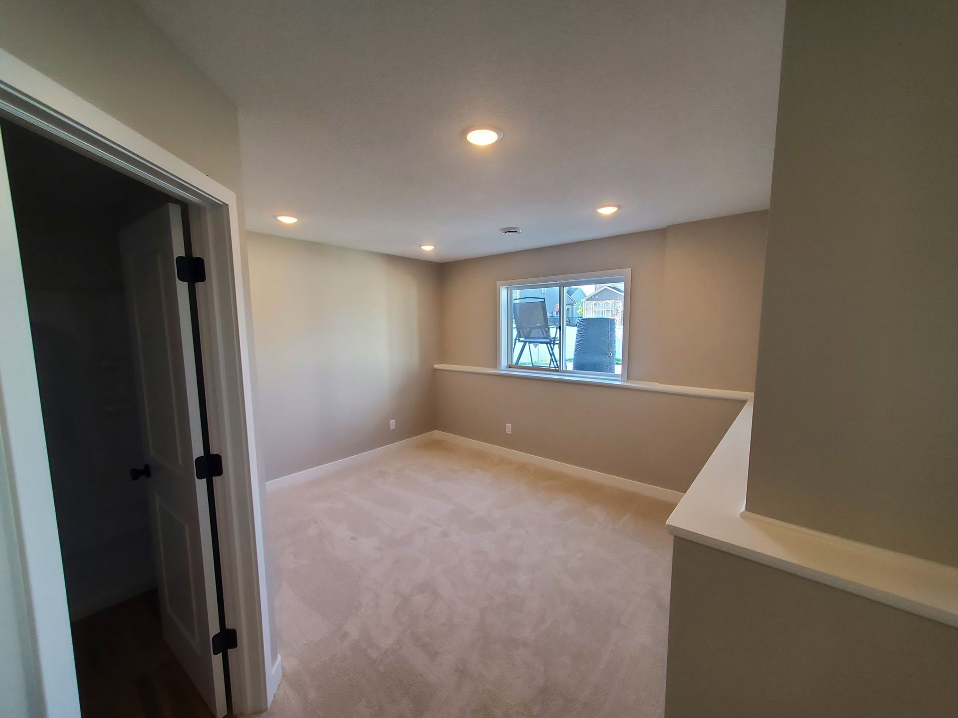 Empty beige room with carpet, recessed lights, and a small window overlooking the street