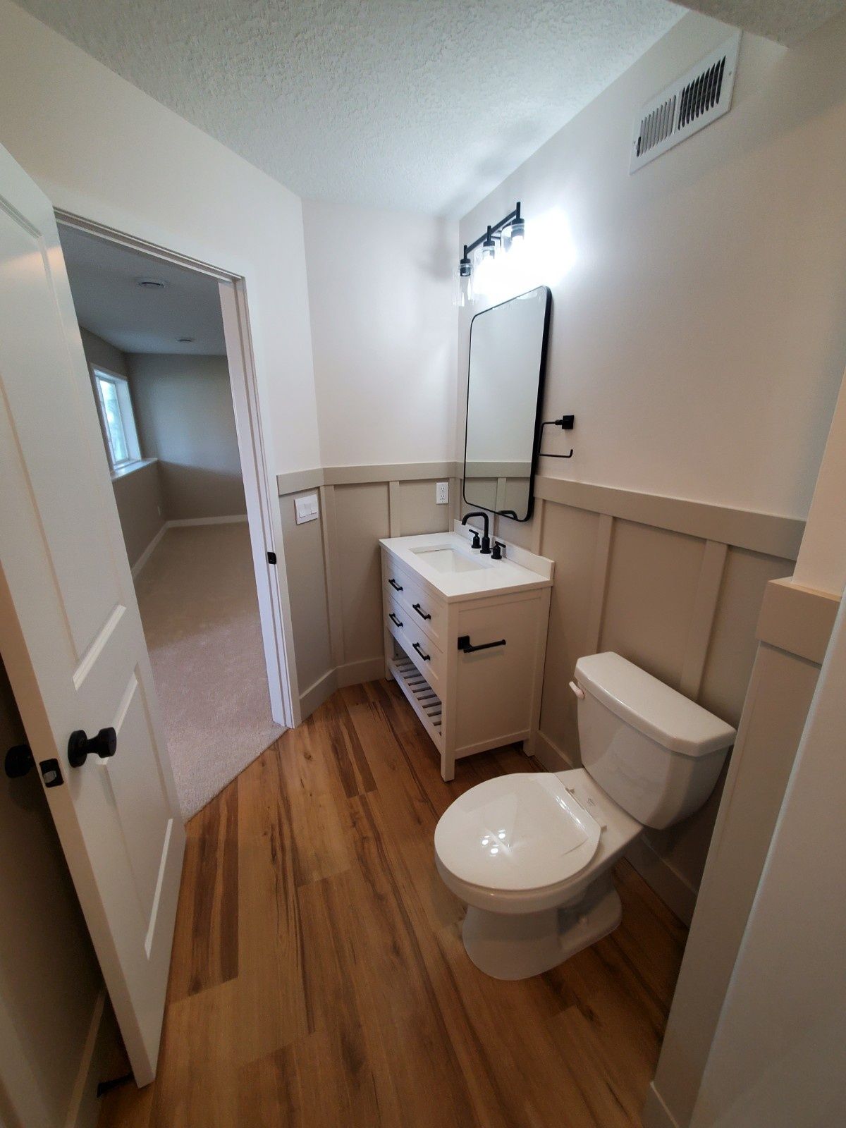 Small bathroom with toilet, vanity sink, mirror, and wood-look floor; open doorway to adjacent room.