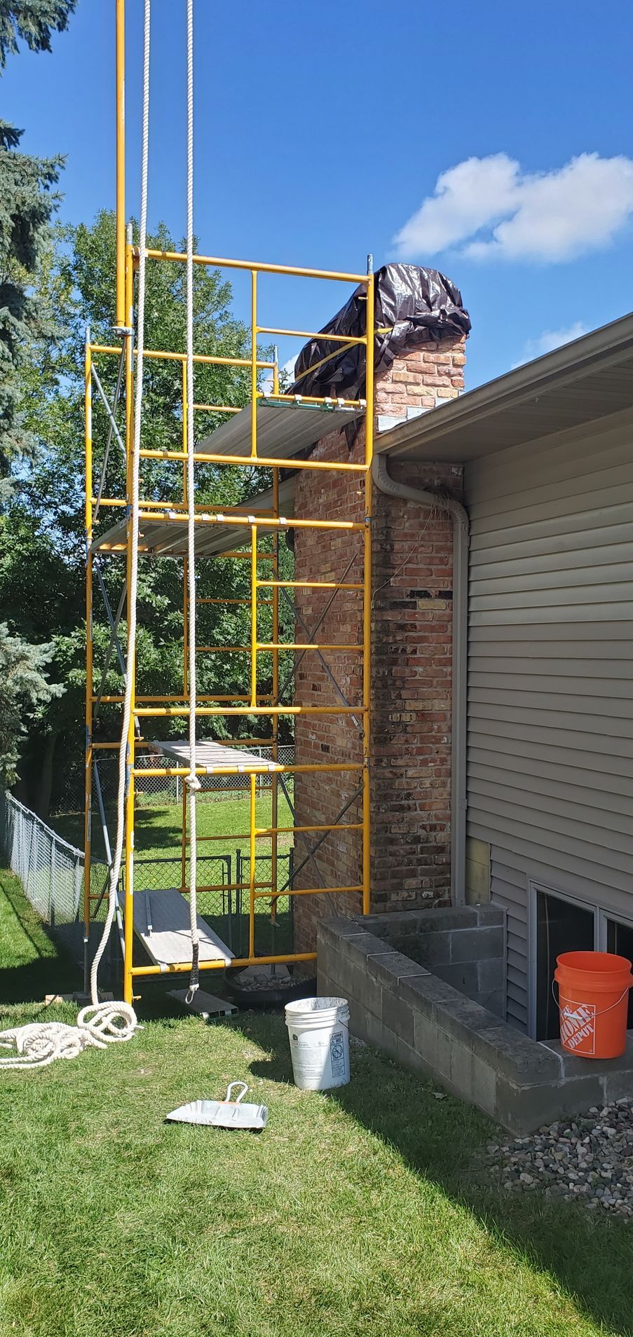 Worker on yellow scaffold outside a brick house beside a white siding wall under a blue sky