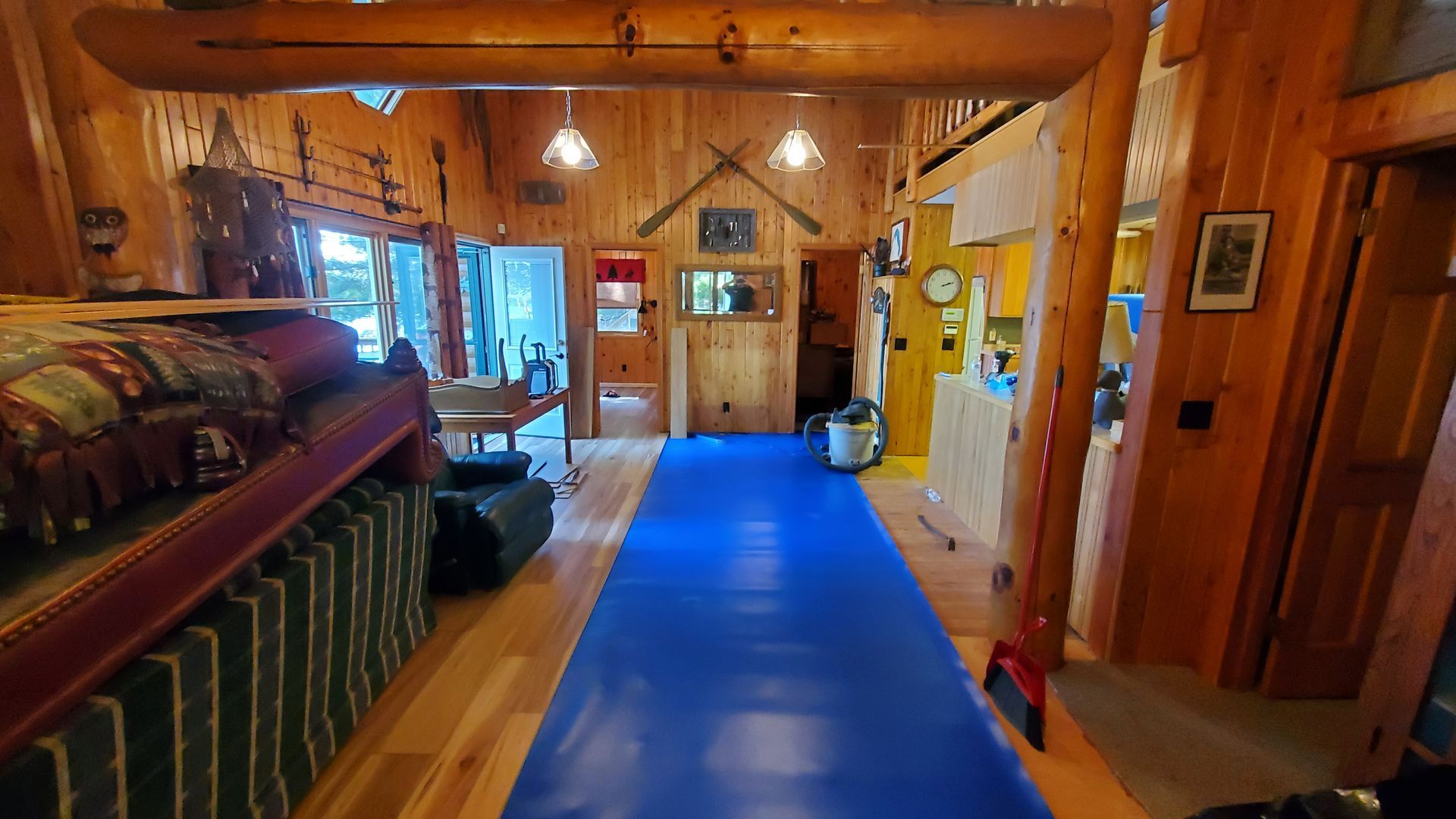 Rustic cabin interior with wooden walls, a blue runner, and a dining area visible through the doorway