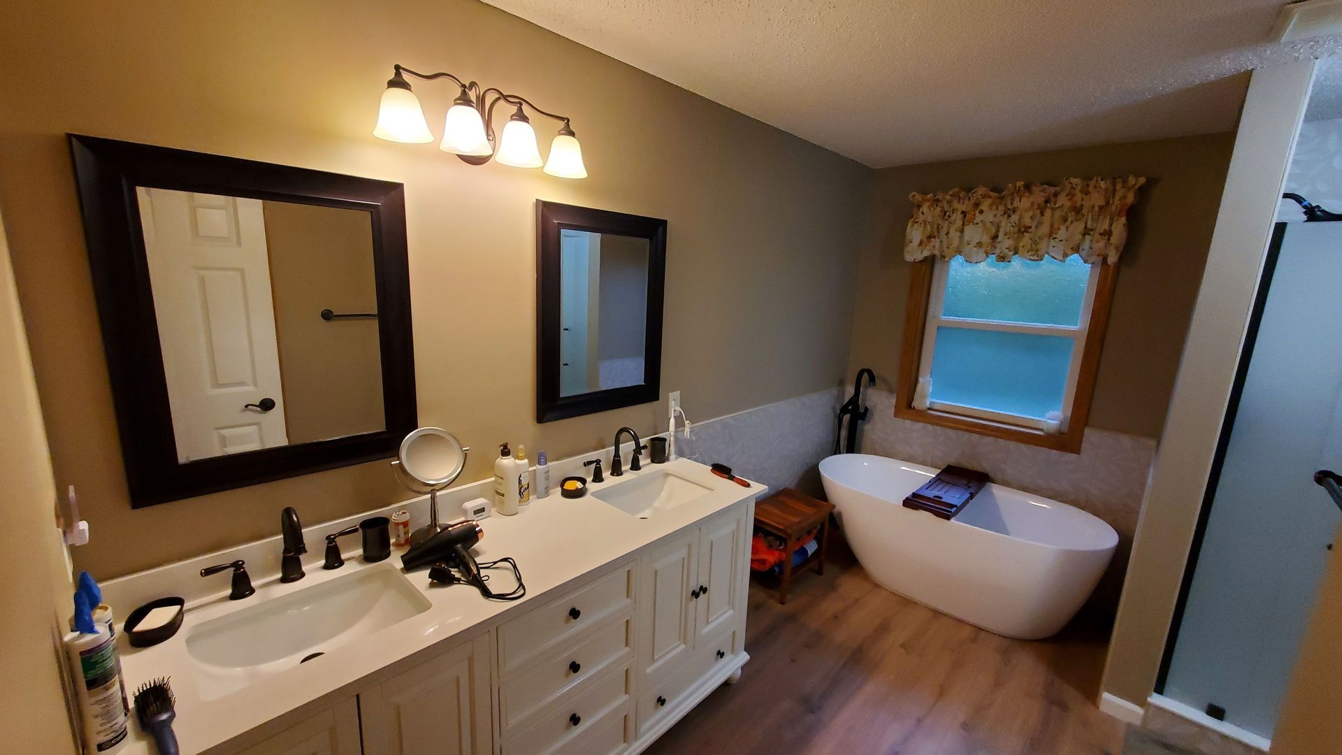 Bright bathroom with dual vanity, mirrors, and a freestanding tub by a window with a valance.